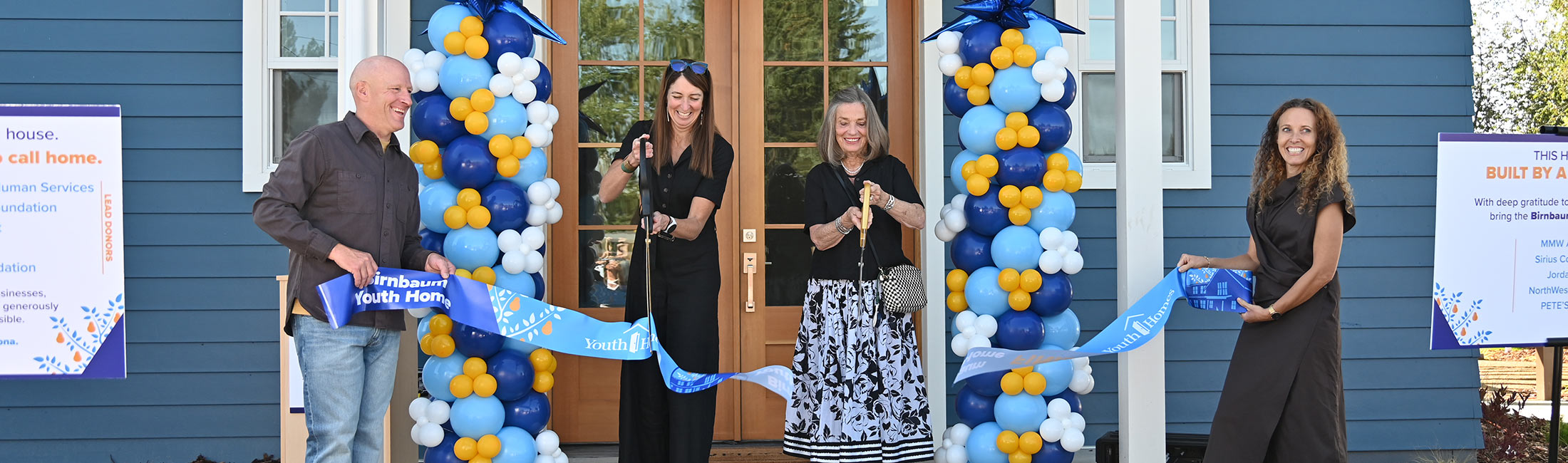 Four people hold a blue ribbon outside the Birnbaum Youth Home entrance as two women in the center cut it with oversized scissors, surrounded by blue, light-blue, white, and yellow balloon columns.