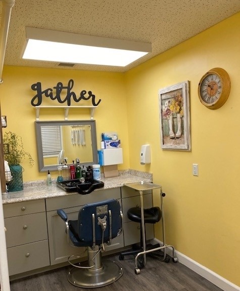 A picture of the salon at the facility. There is a chair, a vanity mirror and counter, and the walls are yellow. There is a sign that says “gather” above the mirror.