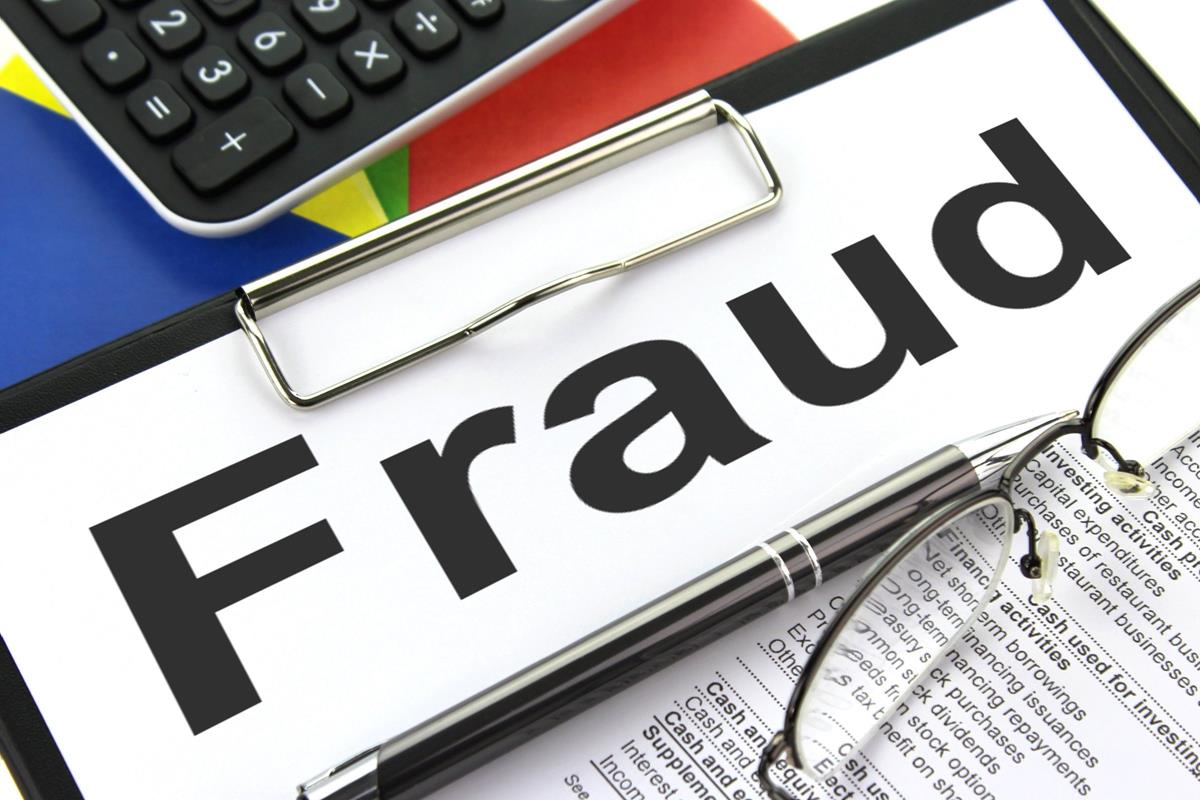 Photo of Paper with the word "Fraud" clipped to a clipboard, with part of a computer keyboard and reading glasses showing